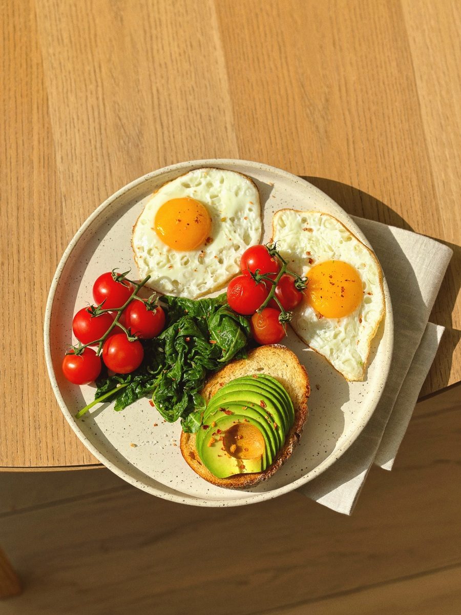 Overhead breakfast in bright sun: two fried eggs, cherry tomatoes on the vine, spinach and avocado toast