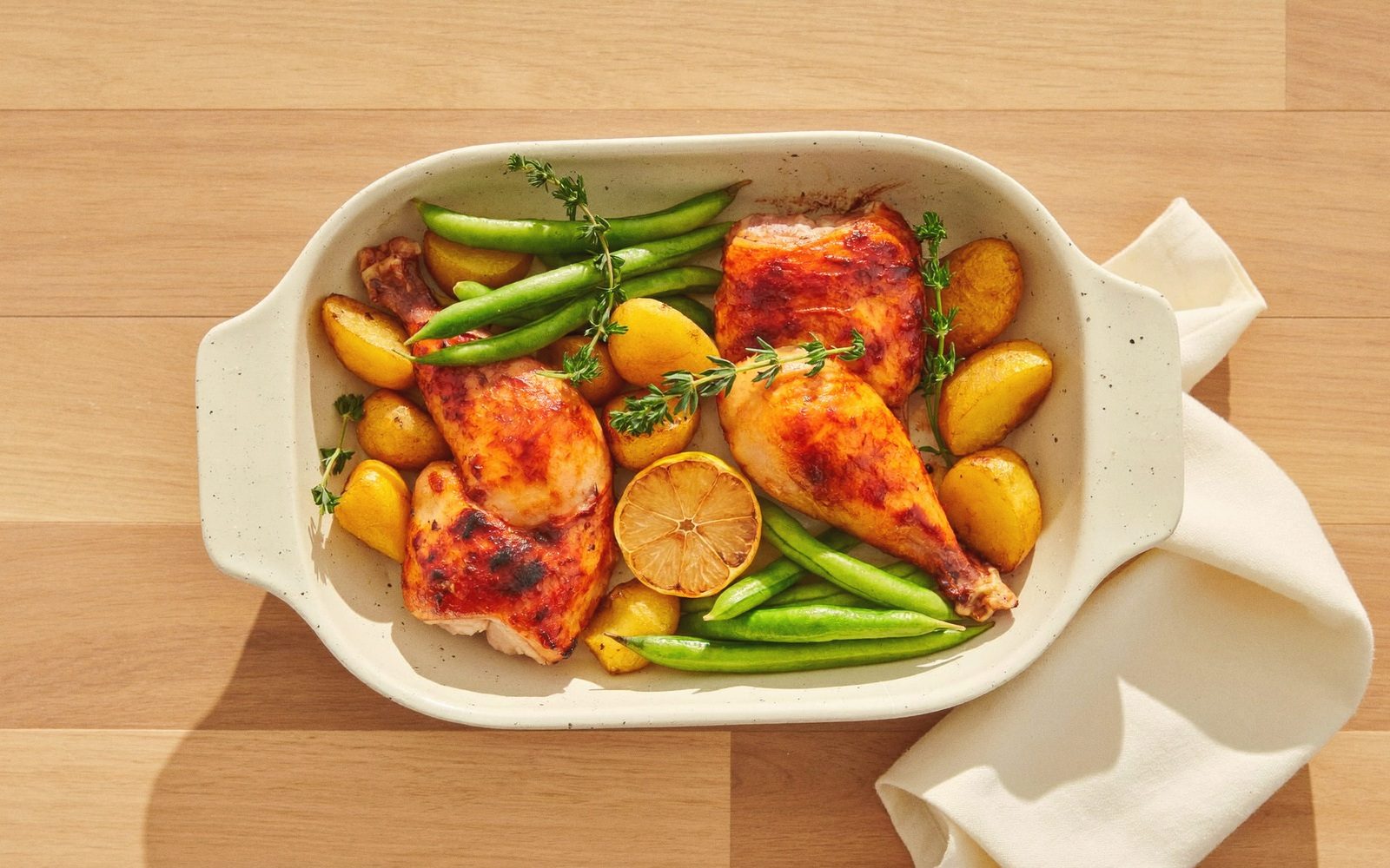 Overhead roast chicken legs with baby potatoes, snap peas and a roasted lemon in a white baking dish
